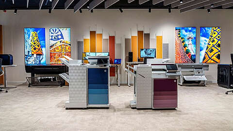 Zwei moderne Multifunktionsdrucker stehen in einem gut beleuchteten Ausstellungsraum mit bunten Architekturfotos an den Wänden und Designelementen wie Akustikplatten und Computerarbeitsplätzen im Hintergrund.