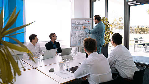 Fünf Männer in Geschäftskleidung sitzen um einen Konferenztisch mit Laptops und Papieren, während ein Mann in einem hellen, modernen Büro mit großen Fenstern steht und Informationen auf einem Whiteboard präsentiert.