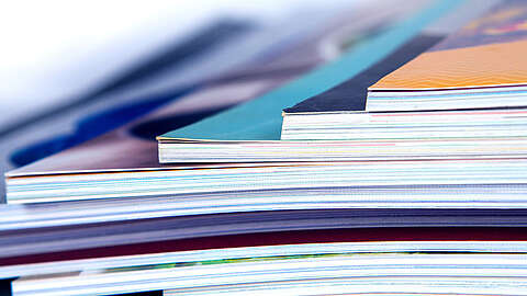 Nahaufnahme der Ränder eines Stapels von Zeitschriften oder Katalogen, die bunte Einbände und sauber ausgerichtete Seiten zeigen. Das Foto unterstreicht das mehrschichtige, strukturierte Aussehen der gestapelten Publikationen.