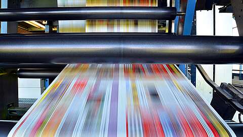 Nahaufnahme einer sich schnell bewegenden industriellen Druckmaschine, in der bunte Papierbögen schnell durch große schwarze Walzen laufen. Die gedruckten Bilder erscheinen aufgrund der Geschwindigkeit der Maschine unscharf.