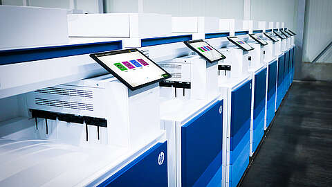 Managed Print Services: Drucker Eine Reihe großer Industriedrucker. Jeder Drucker verfügt über eine Touchscreen-Oberfläche mit bunten Grafiken. Die Drucker sind überwiegend weiß mit blauen Verkleidungen. Der Boden besteht aus grauem Beton.