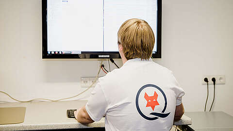 Eine Person mit hellbraunen Haaren, die ein weißes Hemd mit einem roten Fuchs-Logo auf dem Rücken trägt, sitzt an einem Schreibtisch vor einem großen Monitor, auf dem Informationen angezeigt werden, in einem hellen, modernen Büro.