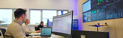 Drei Personen arbeiten an Computerschreibtischen in einem modernen Büro mit großen Monitoren, die Daten und Grafiken an der Wand anzeigen. Eine Person konzentriert sich auf einen gebogenen Bildschirm, und durch die Fenster im Hintergrund fällt natürliches Licht ein.