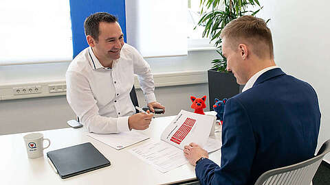 Fleet Management durch Managed Print Services Zwei Männer in Geschäftskleidung sitzen sich an einem Schreibtisch gegenüber, lächeln und besprechen Dokumente. Ein Mann hält ein Papier mit Diagrammen in der Hand; auf dem Tisch stehen Tassen, Papierkram und ein Laptop. Zur Büroausstattung gehören Pflanzen und Plüschtiere.
