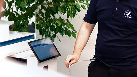 Eine Person in einem marineblauen Hemd benutzt eine Zugangskarte und einen ausziehbaren Ausweishalter an einem elektronischen Kiosk, wobei im Hintergrund eine grüne Pflanze und ein digitaler Bildschirm zu sehen sind.