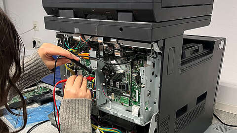 Eine Person testet oder repariert die interne Elektronik eines großen Bürodruckers, dessen Seitenwand geöffnet ist, wodurch Leiterplatten und bunte Drähte sichtbar werden.