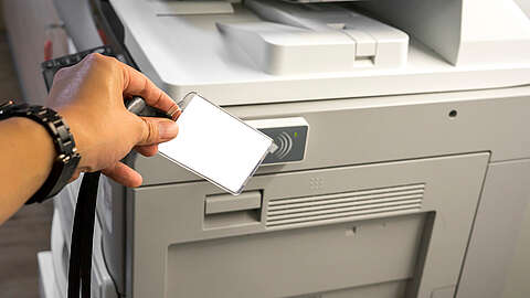 Print Management: Secure Printing Eine Person hält einen Ausweis in die Nähe eines Kartenlesers an einem Drucker/Kopierer, möglicherweise um auf Druckfunktionen zuzugreifen. Der Drucker ist grau und befindet sich in einem Büro.